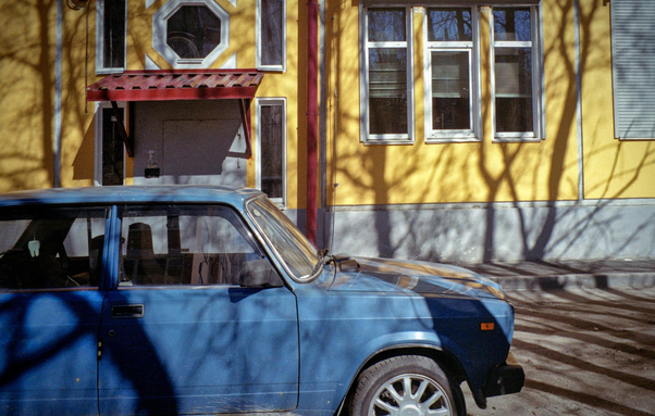There is a blue Lada in the photo, and behind it is a part of a yellow house.