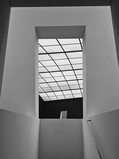 A low-angle, black and white shot of a rectangular opening framed by thick, light-colored walls, leading up to a ceiling composed of many square, translucent panels with a dark grid pattern. Below the ceiling, through the opening, a dark wall is visible, with a small, light-colored rectangular opening in it. A handrail is visible on the bottom right, suggesting a staircase leading to this opening in the foreground.