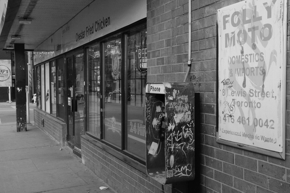 The facade of a corner store recedes from right foreground to left background. The windows are barred against the brick. At far left, a black pillar is stained and had chipped, damaged paint. Centre right, near the bottom of the frame, a payphone has been vandalized and covered with tags and graffiti. At right, a faded, defaced sign advertises a business named Folly Moto.
