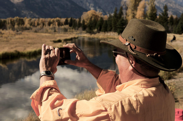 A photographer standing at a river with mirrored, calm water wearing a cowboy hat stretching out his hands clamping a small, chinese made mobile phone with which he is taking likely poor-quality images of the serene, majestic landscape while carrying a slight smirk on his face, as if he had been caught in the action of doing something unlawful, or worse. In the background the river leads towards some trees and mountains further in the back. The whole image has an orange cast to it, maybe because the sun was low, maybe because of poor post processing, maybe because orange is one of the reference colors of the Nikonians community. Hard to tell.