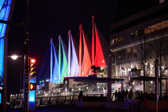 Night time photograph with lots of black and grey tones of some buildings with light inside, people outside. Five tall, colourful flags outside lit from below.
