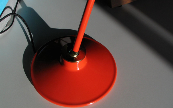 Closeup of a red lamp with a circular base, a spherical-jointed red rod displaying a star-shaped highlight of the sun.