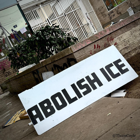 Front of a graffiti tagged I C E building. A large sign with block letters leans against a concrete planter, reads Abolish I C E 