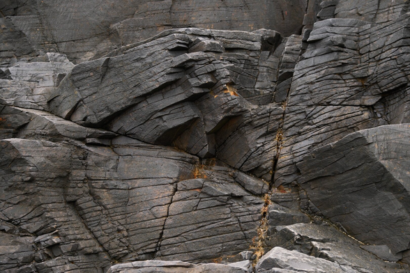 An abstract-ish photo of large sea stack rocks on a beach. The rocks are dark grey with fracture lines running mostly parallel and changing depending on the shape of the rock.