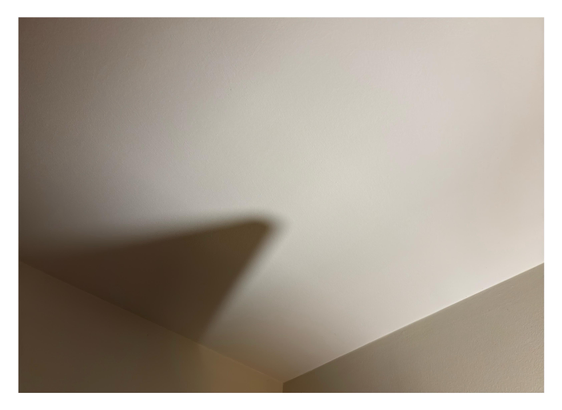 Beige-toned photo of a room interior, looking up at a plain ceiling. The corner where the ceiling and two walls meet is at bottom center, visually forming a low “Y.” On the left side of the image, lamplight throws the shadow of an open door onto the ceiling, appearing as a dark, rightward-tilting triangle on the left arm of the “Y.” The overall effect is minimalistic and abstract.