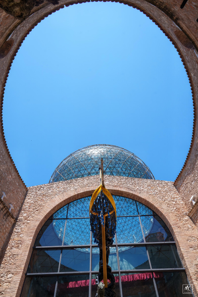 Color photo looking up at a blue sky framed by the ellipse-shaped open top of a building courtyard with a geodesic glass spherical dome and a large arched window at one end of the ellipse. In the center is a the top of a surrealistic sculpture with a boat propped up in the air with large water drops suspended below it. 