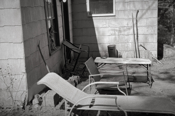 This photograph shows a sunlit, cluttered patio or backyard area next to a house with weathered siding. Several mismatched chairs and a table are scattered around, some tipped over or leaning, with a few cinder blocks on the ground and a barrel or trash can in the corner. A doll or mannequin part is visible in the window, adding an eerie detail to the otherwise abandoned, utilitarian scene.