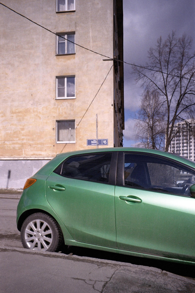 The photo shows a green car parked next to a multi-storey ochre house.