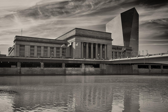 A large neoclassical train station across a river. To the right of the station is a modernist glass office tower.