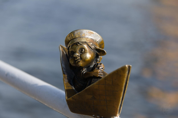 The image captures a close-up view of a palm-size bronze sculpture featuring the head and shoulders of a boy sitting in a paper ship. He wears sailor's striped vest and peakless hat wit his drooping long ears clearly visible. His head turned to the left side of the frame and his hands are wrapped around the central part of the paper ship.

The image is highlighting the natural texture and color variations characteristic of bronze material - ranging from deep browns to rich gold tones that reflect light subtly. The background is blurred but suggests an outdoor setting with hints of water, possibly indicating the statue's location near a river.