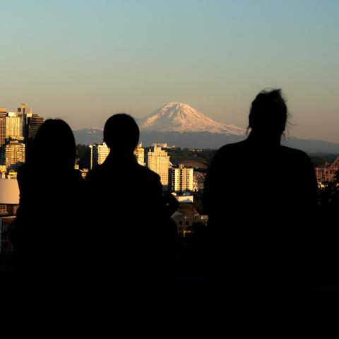 Shot of a family of three, in silhouette, against part of the Seattle skyline. Between the heads of the mother and her two children, Mt Rainier sits in the warm illumination of the setting sun.