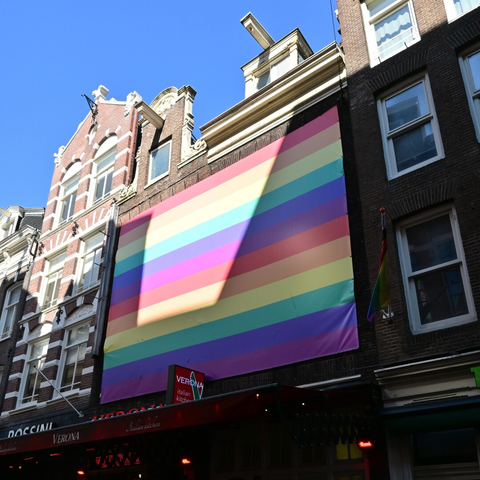 Ein großes Regenbogenbanner an einem historischen Stadthaus, das Sonnenlicht bildet darauf ein Trapez. Unter dem Banner befindet sich ein italienisches Restaurant.