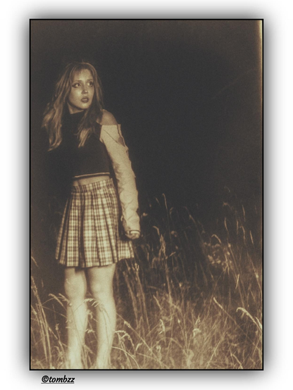 A girl stands in tall grass at night, against the backdrop of a dark, desolate area. Suddenly illuminated by a car’s headlights, she looks terrified — her mouth is open, eyes wide, body tense and slightly turned as if ready to run. She’s wearing a short top and a plaid skirt. Her long, loose hair falls across her face. The entire scene radiates tension, fear, and a sense of sudden danger, like a frame from a thriller. The film photography adds a grainy, moody texture with desaturated tones and deep shadows, evoking a raw, vintage aesthetic that heightens the unease and cinematic atmosphere.