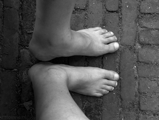 Two bare feet resting on cobblestones. The feet appear to belong to the same person, with one foot positioned above and slightly behind the other. 