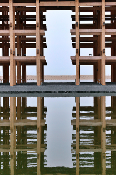 Die gerüstartige Konstruktion aus hellem Holz, spiegelt sich in einem künstlichen See.