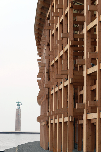 Teil des Außenrings aus miteinander verzahnten Holzbalken.
Davor ein eingefaßtes Stück See, die sich vom Meer abgrenzt. 
Im Hintergrund ein Sendeturm.