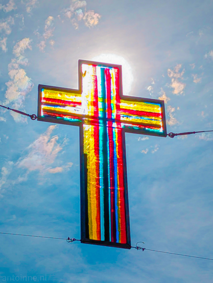 A vibrant, colorful cross, made of stained glass, hanging against a bright, slightly cloudy sky. The sun is positioned directly behind the cross, creating a powerful silhouette effect and causing the colorful panes of the cross to glow intensely. The cross is filled with vertical and horizontal stripes of various colors, including reds, yellows, greens, and blues, which appear to shimmer and blend due to the strong backlight. The sky behind it is a light blue with scattered white, wispy clouds, some of which show subtle iridescence, particularly near the sun's position. The overall impression is one of striking visual contrast and luminous color.

The St. John's procession in the Gooische Laren is a procession in honor of the patron saint St. John. Laren is the only Dutch municipality above the rivers where a Catholic procession is held.
