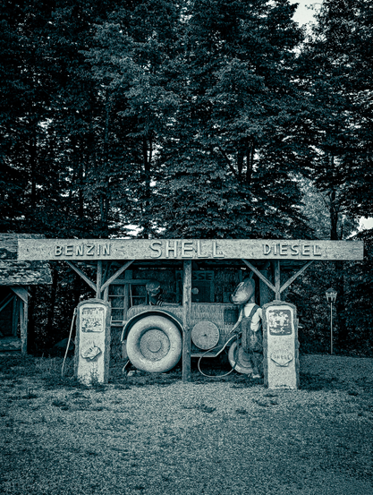 A black-and-white photograph of an old gas station that has been created from straw and wood. Underneath the wooden canopy, there's a central, large, and somewhat whimsical structure that resembles an old tractor, with oversized, textured wheels and various mechanical components. To the left and right of this central structure are two vintage gas pumps, appearing to be made of concrete or stone, with old-fashioned gauges and nozzles. They have "SHELL" logos visible. Above the entire setup, a prominent sign spans the width of the structure, bearing the words "BENZIN SHELL DIESEL".