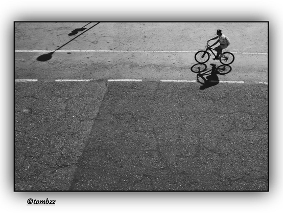 Black-and-white minimalist photograph featuring a woman riding a bicycle in the top right corner of the frame. Her shadow is clearly cast onto the asphalt road surface. In the top left corner, shadows of road signs are visible. White lines run across the asphalt: one solid and one dashed. The composition is enhanced by the sharp sunlight of a clear day, emphasizing contrasts and lending the image a strong graphic quality.