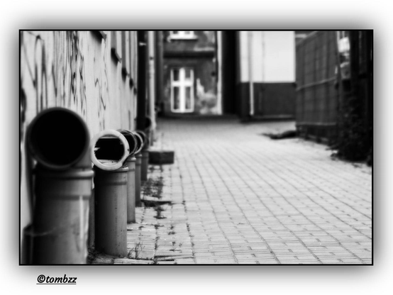 A black-and-white photograph shows a narrow, enclosed courtyard between tall tenement buildings. On the left side of the frame, massive metal drainpipes run down — dominant in the composition, as if frozen mid-scream, echoing in the dead silence of the architecture. Next to them, a wall with rectangular windows, some open. To the right, the wall of another building is visible. In the background, through a gap between the walls, a closed gate leading to the street can be seen, with a single window beside it. The scene carries a sense of tension and symbolism, where technical elements — like the pipes — take over the emotional narrative.