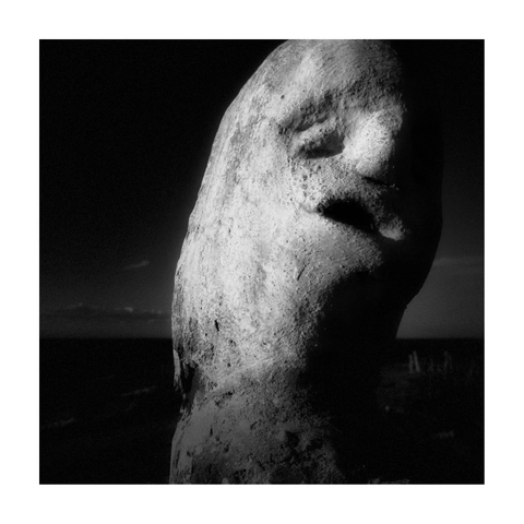A square black-and-white photograph framed with a white border, showing a close-up of a weathered sculpture with humanoid features, positioned near the sea. The figure fills the right side of the frame and is side-lit, casting deep shadows that accentuate its rough, eroded surface. Its face appears to bear empty eye sockets and a distorted mouth frozen in a silent scream, giving the impression of pain or anguish. The dark background, with a faint horizon and subtle hints of ocean, reinforces the sense of isolation. Strong grain and high contrast contribute to the raw, haunting atmosphere.