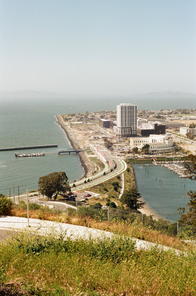 A lone, tall building on the coast line of Treasure Island, with the road up Yerba Buena Island in the foreground. 