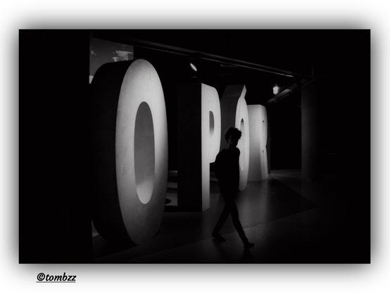Black-and-white photograph taken in the dimly lit exhibition space of the Museum of the Second World War in Gdańsk. In the foreground, a young man dressed in dark clothing is captured in motion—his figure sharply stands out against the shadowy background. Behind him, large three-dimensional letters spell out the word OPÓR (RESISTANCE), forming part of the exhibition. The composition and interplay of light give the image a dramatic, almost cinematic atmosphere.