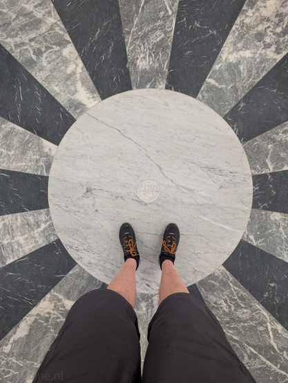 Looking down at a person's feet standing on a circular marble inlay. The person is wearing black shorts and dark athletic shoes with orange laces. The marble inlay features a central light grey or white circular disc. Around this central disc, there's a pattern of alternating dark grey/black and light grey/white trapezoidal segments radiating outwards, creating a sunburst effect. In the very center of the light grey disc, there's an emblem or marking that appears to include the numbers "1”, “9", "8” and”3".