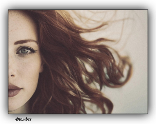 Portrait of a young woman captured in an artistic close-up. Half of her face is visible, giving the composition an intriguing character. Her chestnut hair is dynamically windswept, creating a sense of movement and lightness. Intense makeup highlights her gaze – her eyes are emphasized with a bold line and thick mascara, drawing attention. Her lips are coated in a deep, saturated lipstick, adding sensuality and strength to the image. The overall effect radiates elegance and a deliberate sense of aesthetics.