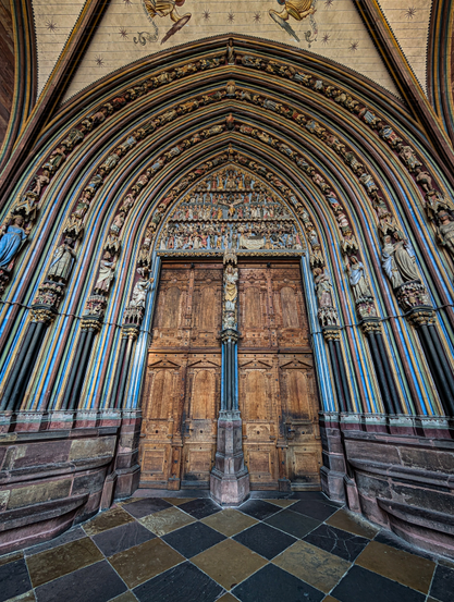A low-angle, wide-shot photograph of a grand, ornate Gothic cathedral entrance. The focal point is a massive double wooden door, aged and weathered, with intricate carvings on its upper panels. Above the doors, the archway is deeply recessed and features multiple layers of detailed sculptures and architectural elements. Each arch is adorned with rows of figures and narrative scenes, characteristic of medieval religious art. 