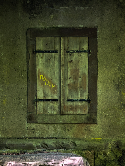 A rustic, closed wooden window set into a weathered stone wall. The window features two vertical wooden shutters, each secured by two black, wrought-iron hinges. The wood of the shutters is a muted, desaturated green or olive tone, showing signs of age and exposure to the elements, possibly with some moss or discoloration. A horizontal line across the middle of the shutters suggests where they meet when closed. Prominently spray-painted in bright yellow on the left shutter are some letters. The window is framed by a darker, possibly stone, lintel and sill, which are slightly recessed from the main wall. The surrounding wall is textured and appears to be made of rough-hewn stone, also discolored with patches of green and dark areas.
