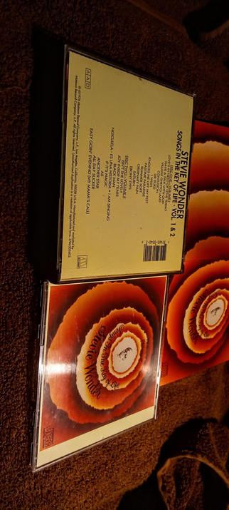 The photograph shows an open CD case with a vibrant, concentric circular design in shades of orange, red, and yellow. The design features a gradient effect, with the colors transitioning smoothly from the center to the edges. The text "Songs In The Key of Life" is prominently displayed in a cursive font within the central circle. The CD case is labeled "Compact Disc Digital Audio" and includes a barcode and copyright information. The CD case is placed on a brown carpeted surface, and the lighting casts a warm glow on the design, enhancing its visual appeal.