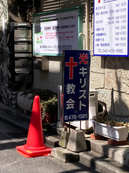 A red traffic cone stands beside a blue sign with a white cross and Japanese text indicating a church. In the background, there are mailboxes and a notice board with various announcements. The setting appears to be an urban area with a paved walkway.