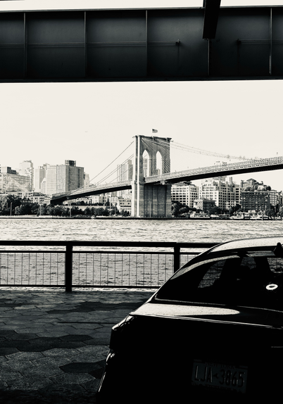 A car parked under an overpass by the East River. Across the river is the Brooklyn Bridge 