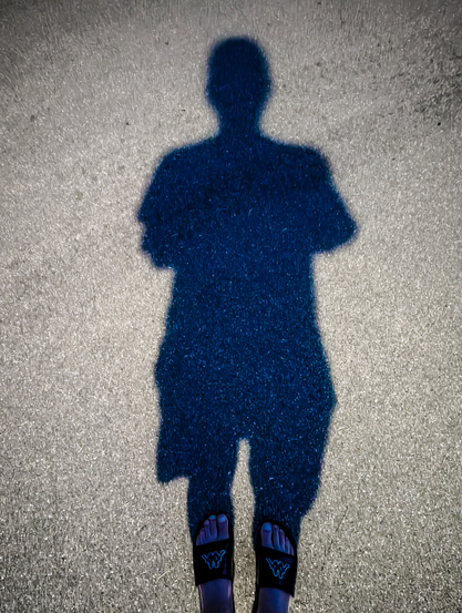 The shadow of a person cast onto asphalt. The shadow is clearly defined, and shows the full upper body of a person wearing a t-shirt and shorts. The person's feet are visible at the bottom of the frame, wearing dark flip-flops with a white Kappa logo on each. The ground around the shadow is brightly lit by a street light and shows a coarse, grainy texture.