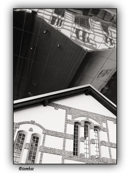Black-and-white photograph capturing a striking architectural contrast: a historic building with tall arched windows and a steeply pitched roof is reflected in the glass façade of a modern structure that looms above it with unsettling lightness. The geometric order of contemporary architecture seems to absorb the subtle elegance of the past, creating a visual dialogue between eras.