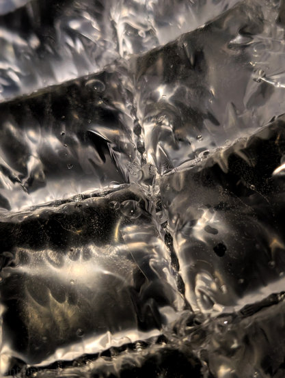This is a close-up, abstract photograph that captures a dynamic and somewhat chaotic scene of water interacting with a textured and rippled surface, made of plastic.

The image is dominated by various shades of silver, grey, and black, with glints of light creating highlights that suggest reflectivity. There's a strong sense of movement, with swirling patterns and what look like bubbles or air pockets suspended within the liquid. Some areas are sharply defined, showing distinct lines and creases, while others are blurred, contributing to the sense of fluidity. 