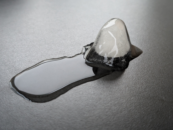Close-up, high-angle shot of a melting ice cube on a dark, textured surface. The ice cube itself is partially translucent, with some cloudy white areas indicating air bubbles or impurities within its structure. It's an irregular, somewhat conical shape, with its base already melted into a small puddle. 

The puddle of water extends from the base of the ice cube in a narrow, elongated stream towards the bottom left of the frame. The surface of the water is reflective, showing some subtle highlights from an unseen light source. The dark surface beneath the water and ice creates a strong contrast, making the clear water and the ice cube stand out. 