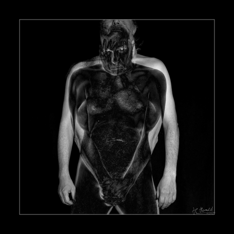 Square picture with black frame and black background. A naked man stands facing the viewer. His arms hang loosely at his sides. You can see him from nose to thigh. In front of him, or rather inside him, is another black figure who is protecting its nakedness with his hands and holding his hands in front of its head at the same time. The black figure looks into the camera with lucid eyes and keeps its head lowered. 
The picture is a composition of three different shots, which are superimposed and partly inverted.