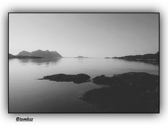 A black-and-white landscape photograph capturing a tranquil scene of nature. In the foreground, dark, jagged rocks emerge from a smooth water surface, adding depth and contrast. The water reflects the diffused glow of the sky. In the background, blurred outlines of mountains and islands stretch across the horizon, forming a serene silhouette. The entire composition evokes stillness and quietude.