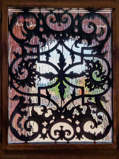 An ornate, dark wooden grille set within a window frame, viewed from the interior. The grille features intricate, swirling, and symmetrical patterns, including scrollwork and botanical-like motifs, particularly a central, almost cruciform design with leaves. The glass itself seems to have a slightly textured or wavy quality, creating a subtle distortion of the light and the view beyond. The entire window is encased in a dark wooden frame, which is visible on all four sides of the image. 