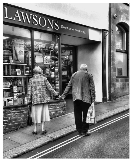An elderly couple walk hand in hand along the high street. The wife is looking into an interesting shop window while the husband looks on. The most beautiful discovery true friends make is that they can grow separately without growing apart. As photographed in Totnes, UK