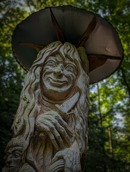 A wooden sculpture, carved from a tree trunk, set against a blurred background of a wooded area. The sculpture depicts the upper body of an elderly woman, or perhaps a witch-like figure, with intricately carved details. Her face is prominent, featuring deep-set eyes, a wide, somewhat sinister smile revealing carved teeth, and a long nose. She is holding a knife in her right hand, positioned across her chest. Below her right hand, part of another face or figure can be glimpsed, adding to the mysterious nature of the artwork. Above her head, there's a large, round, dark object that resembles an upside-down mushroom cap, providing a sort of canopy or roof over the figure. 