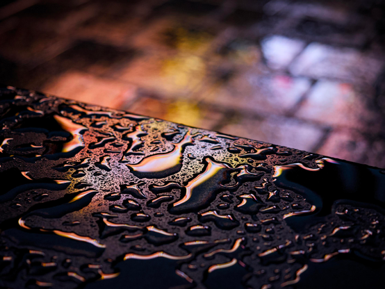 Rainwater pools on the black marble surface of a low bench in St Enoch Square, reflecting a golden neon light from an LED advertising panel high on a shopping centre