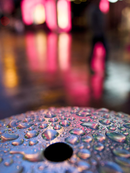 Droplets of rainwater settled on a shiny silver bollard pick up hints of the neon shop frontages on Buchanan Street and a shallow depth of field creates the blurry pink background and the outline of a passing stranger