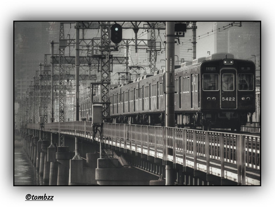 The image depicts a train crossing a railway bridge. The composition is rendered in black and white, lending the frame a nostalgic, documentary-like atmosphere. Train number 5422 moves along steel tracks supported by massive concrete pillars. Above the train hangs a signal light glowing green, indicating permission to proceed. In the background, dense railway infrastructure unfolds with metal poles, electric cables, and structural supports, forming a network of industrial geometry. The frame is carefully balanced—the motion of the train contrasts with the stillness of its surroundings.