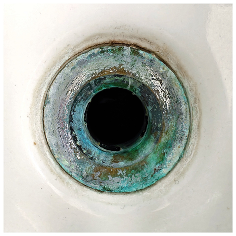 A circular black hole in white ceramic sink, with green-blue copper oxidation stains around the rim. 