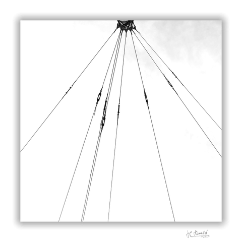 Square picture with white frame. The top of a thick mast can just be seen at the top edge, to which 8 wires are attached for tensioning tram overhead lines. The wires run downwards in a fan shape and each has an integrated tensioning device. The sky is white except for a little grey in the top right-hand corner.