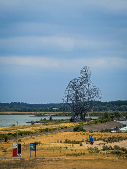 This photograph captures a large-scale outdoor sculpture set against a scenic waterscape under a bright, cloudy sky. The dominant feature of the image is the massive sculpture, made of metal, a complex, open-framework representation of a crouching or sitting human figure. The sculpture is positioned on an artificial embankment separating a body of water from a foreground area.

Exposure is a steel frame sculpture in Lelystad, Netherlands, by artist Antony Gormley. The work is also known as Crouching man (Squatting man), or popularly called the Shitting Man. It is located on the Markerstrekdam near the Houtribsluizen.