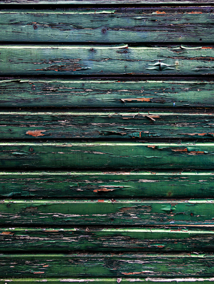 A weathered wooden wall, characterized by horizontal planks. The dominant color is a faded and peeling green paint, revealing patches of the lighter, natural wood beneath. The paint shows significant signs of wear and tear, with numerous chips, cracks, and flakes exposing the lighter-toned wood. 